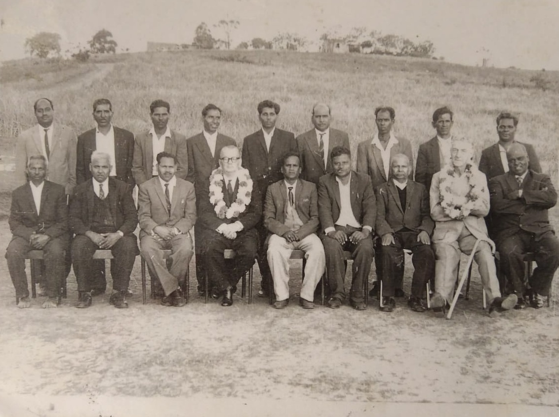 A formal black and white group portrait of eighteen men in suits, seated and standing in two rows on open ground
