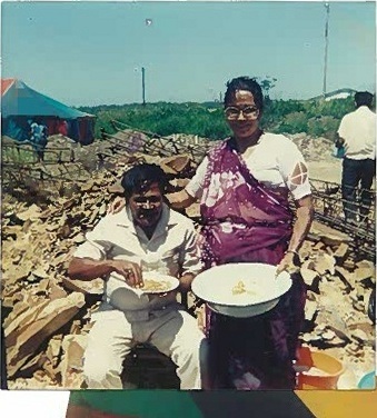A colour photograph of a woman in a sari holding a dish beside a seated man on stony ground, with a marquee behind them