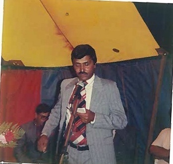 A colour photograph of a man in a suit speaking at a microphone beneath a striped marquee