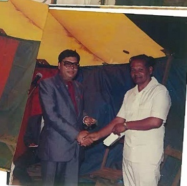 A colour photograph of two men shaking hands beneath a marquee, one handing the other an envelope