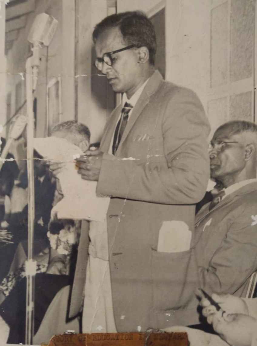 A man in a suit and glasses standing at a microphone, reading from a sheet of paper, with seated guests beside him