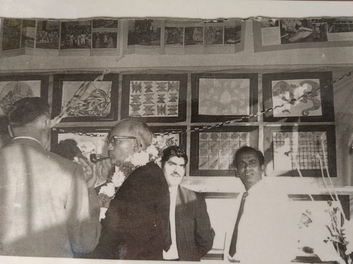 A black and white photograph of men gathered beneath a wall of framed artwork, one wearing a garland and holding a pipe